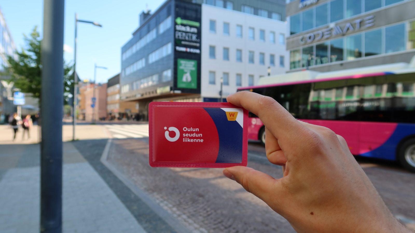 Henkilöllä kädessä Oulun seudun liikenteen matkakortti. Taustalla ajaa ohitse linja-auto.