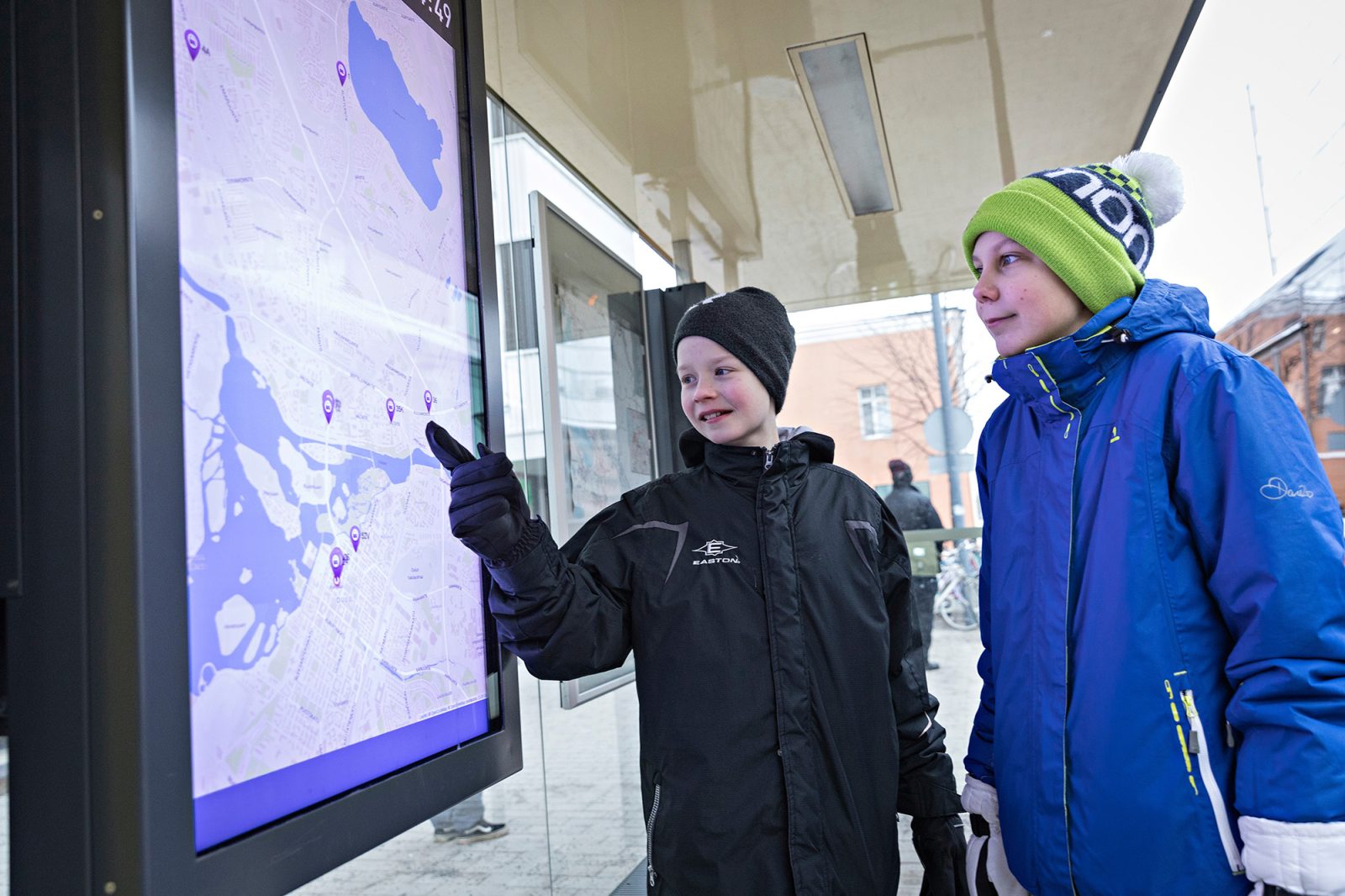 Kaksi poikaa bussipysäkillä diginäytön karttaa katselemassa talvella.
