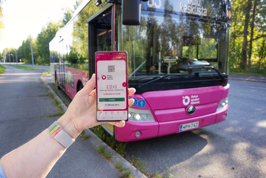 Puhelimen näytössä mobiililippu ja bussi taustalla pysäkillä.